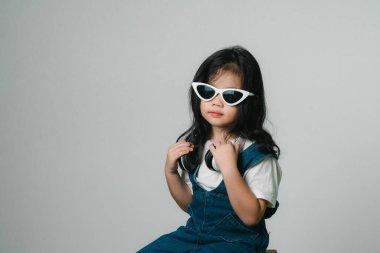 Rahatlamış, sevimli Asyalı kız çocuğu portresi. Gülen, gülen, beyaz kulaklık takan, elleri kulaklarında, poz veren, gülümseyen, gri arka planda izole edilmiş, mutlu bir şekilde..