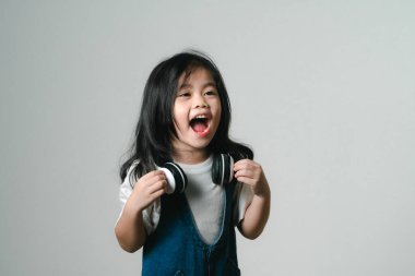 Rahatlamış, şirin Asyalı kız çocuğu portresi. Gülen, gülen, beyaz kulaklık takan, elleri kulaklarında, poz verip gülümseyen, gri arka planda izole edilmiş..