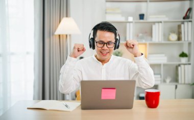 Asyalı iş adamı gözlük takıyor ve kulaklık takıyor. Dizüstü bilgisayar üzerinde çalışırken ve evde kutlama yaparken başarıyla gülümsüyor. Çalışma başarısı neşe ve mutluluk, iş konsepti.