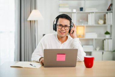 Asyalı bağımsız iş adamı kulaklık takıyor ev telefonuyla video görüşmesi yapıyor, dizüstü bilgisayar kullanıyor, konuşuyor, webinar izliyor ya da dil öğreniyor, konuşuyor, çevrimiçi eğitim veriyor, e-öğreniyor..