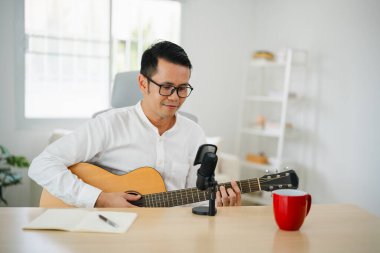 Asyalı adam youtuber canlı yayında gitar çalıyor ve şarkı söylüyor. Asyalı adam gitar öğretiyor ve internette şarkı söylüyor. Dizüstü bilgisayarlı ve akustik gitar çalan müzisyen müziği.