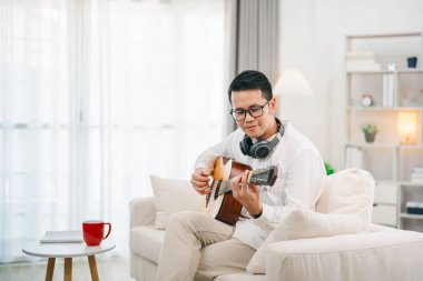 Evdeki oturma odasında kanepede otururken gitar çalan gözlüklü ve kulaklıklı bir Asyalı. Asyalı adam evde gitar çalarken şarkı yazıyor. Şarkı müziği konsepti oluştur.