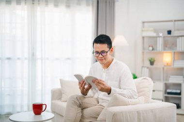 Neşeli Asyalı adam kitap okuyor ve evdeki oturma odasında kanepede otururken sıcak kahve içiyor. Asyalı adam evdeki kanepede kitap okuyup gülümsüyor. Ev kavramında etkinlik hobisi.