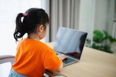 Arka manzara. Turuncu t-shirt giyen Asyalı kız bebek evdeki oturma odasındaki ahşap masada dizüstü bilgisayar ve e-öğrenme kullanıyor. Eğitim öğrenimi online olarak ev kavramından.