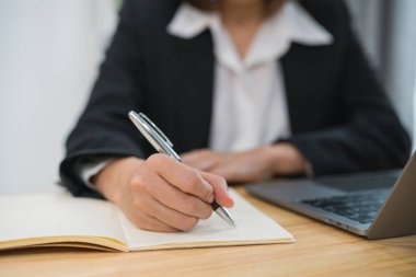 Takım elbiseli, not defteri yazan ve evdeki masada dizüstü bilgisayar kullanan Asyalı iş kadınına yakın çekim. Girişimci kadın iş için oturma odasında çalışıyor. İş yeri ev kavramı.