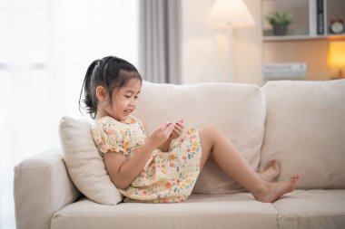Kanepede cep telefonu ekranı kullanan ve dokunan Asyalı kız çocuğu. Bebek gülümsemesi cep telefonu kullanmak için komik bir zaman. Çok fazla ekran zamanı. TV ve internet bağımlılığı varken video izleyen tatlı bir kız..