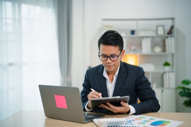 Asyalı iş adamı kalemle not defteri kullanarak gülümsüyor ve ev ofisindeki ahşap masada dizüstü bilgisayarla çalışıyor. Girişimci adam evde iş için çalışıyor. Ev konseptinde iş çalışması.