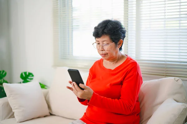 Happy senior old asian woman enjoying using mobile apps texting typing messages sit on sofa, smiling old lady holding smartphone looking smartphone browsing social media or learning technology at home