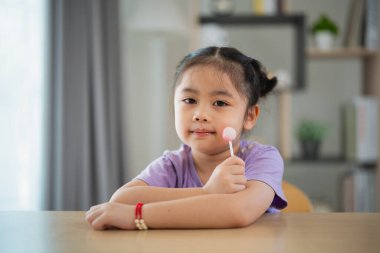 Sevimli Asya 'lı tatlı kız renkli çocuk lolipoplarıyla evdeki oturma odasındaki tahta masada oynuyor ve yiyor..