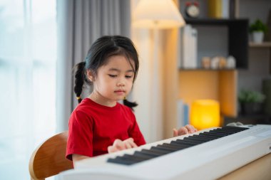 Evdeki oturma odasında piyano çalmayı öğrenen mutlu Asyalı küçük tatlı kız gülüşü. Karantina sırasında çocuk için evde etkinlik fikri. Müzik öğrenme çalışması.