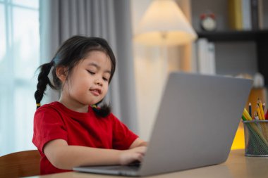Kırmızı tişört giyen Asyalı bir kız bebek evdeki oturma odasındaki ahşap masada dizüstü bilgisayar kullanıyor ve ders çalışıyor. Eğitim öğrenimi online olarak ev kavramından.
