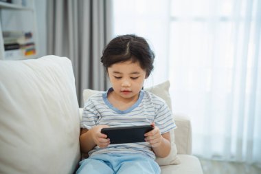 Kanepede cep telefonu ekranı kullanan ve dokunan Asyalı kız çocuğu. Bebek gülümsemesi cep telefonu kullanmak için komik bir zaman. Çok fazla ekran zamanı. TV ve internet bağımlılığı varken video izleyen tatlı bir kız..