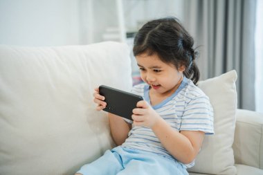 Kanepede cep telefonu ekranı kullanan ve dokunan Asyalı kız çocuğu. Bebek gülümsemesi cep telefonu kullanmak için komik bir zaman. Çok fazla ekran zamanı. TV ve internet bağımlılığı varken video izleyen tatlı bir kız..