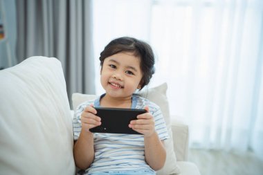 Kanepede cep telefonu ekranı kullanan ve dokunan Asyalı kız çocuğu. Bebek gülümsemesi cep telefonu kullanmak için komik bir zaman. Çok fazla ekran zamanı. TV ve internet bağımlılığı varken video izleyen tatlı bir kız..
