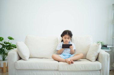 Kanepede cep telefonu ekranı kullanan ve dokunan Asyalı kız çocuğu. Bebek gülümsemesi cep telefonu kullanmak için komik bir zaman. Çok fazla ekran zamanı. TV ve internet bağımlılığı varken video izleyen tatlı bir kız..
