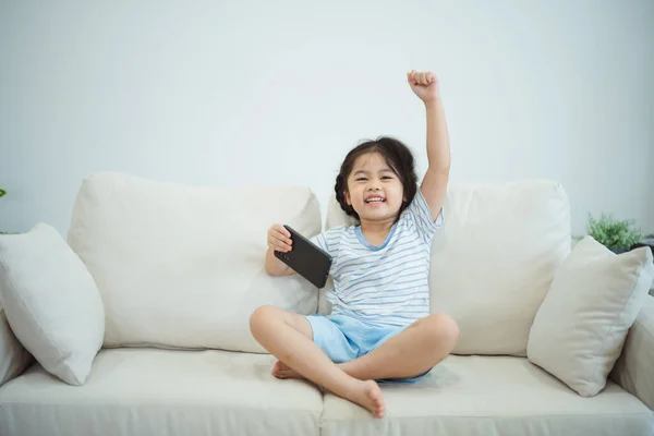 Asian child girl raise your hand cheerful success looking using and touch mobile phone screen on couch sofa. Baby smiling funny time mobile phone. Too much screen time. Internet addiction.