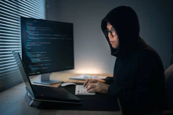 Hacker man wearing hood writing code to hack Network security system. Dangerous Hooded Hacker Breaks into Government Data Servers and Infects Their System with a Virus. Hacking and malware concept.