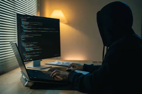 Hacker man wearing hood writing code to hack Network security system. Dangerous Hooded Hacker Breaks into Government Data Servers and Infects Their System with a Virus. Hacking and malware concept.