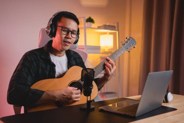 Asyalı adam youtuber canlı yayında gitar çalıyor ve şarkı söylüyor. Asyalı adam gitar öğretiyor ve internette şarkı söylüyor. Dizüstü bilgisayarlı ve akustik gitar çalan müzisyen müziği.