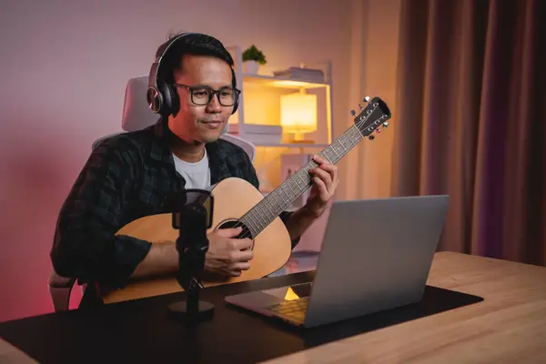 Asian man youtuber live streaming perfomance playing guitar and sing a song. Asian man teaching guitar and singing online. Musician recording music with laptop and playing acoustic guitar.