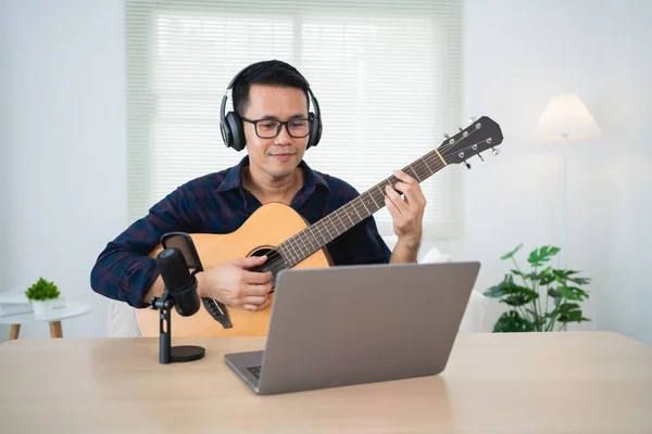 Asian man youtuber live streaming performance playing guitar and sing a song. Asian man teaching guitar and singing online. Musician recording music with laptop and playing acoustic guitar.