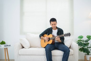 Asyalı bir adam evdeki salonda koltukta ya da yerde gitar çalıyor. Asyalı adam evde gitar çalarken şarkı yazıyor. Şarkı müziği konsepti oluştur.