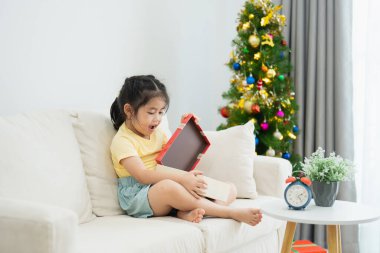 Noel, Noel, kış konsepti. Asyalı küçük kız elinde hediye kutuları, mutlu bir şekilde gülümsüyor. Noel ağacının yanında, evdeki bir oturma odasında yeni yılı kutluyor..