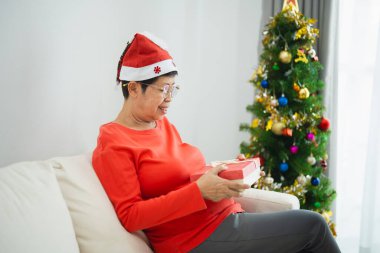 Noel, Noel, kış konsepti. Asyalı yaşlı bir kadın hediye kutularını tutarken mutlu bir şekilde gülümsüyor. Noel ağacının yanında, evdeki bir oturma odasında yeni yılı kutluyor..