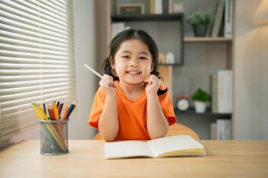 Asyalı kız bebek gülümsüyor. Evdeki oturma odasındaki ahşap masa masasında not defterine renkli kalemler çiziyor. Eğitim öğrenimi online olarak ev kavramından.