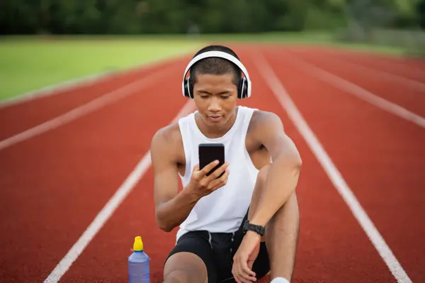 Asian man sportsman fit body wearing white headphone and using mobile phone on white screen to listen music while resting or before jogging, exercising at lane stadium. Sport healthy running concept.