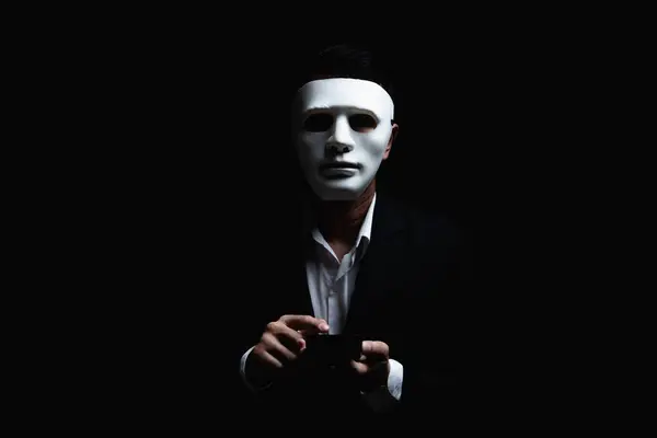 Unknown businessman wearing mask with covered face using mobile phone makes an anonymous call intimidating and threatening the interlocutor on dark background. hacker callcenter concept.