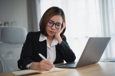 Depresyondaki ciddi Asyalı iş kadını stresli depresyon geçiriyor. Üzücü zaman geçiriyor. Laptop üzerinde çalışıyor ve ofis masasının üzerine not defteri yazıyor. Bunalımlı kadın üzgün ciddi çalışıyor.