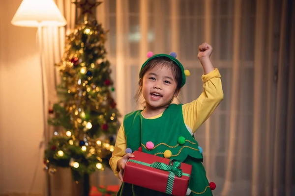 Noel, Noel, kış konsepti. Asyalı küçük kız elinde hediye kutuları, mutlu bir şekilde gülümsüyor. Noel ağacının yanında, evdeki bir oturma odasında yeni yılı kutluyor..