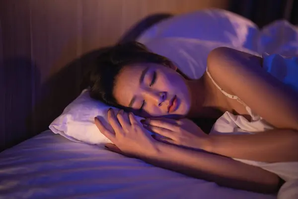 Asian woman sleeping in comfortable bed with silky linens at night light. Women lying in bed and keeping eyes closed while covered with blanket. Content and peaceful, cheerful and comfortable.