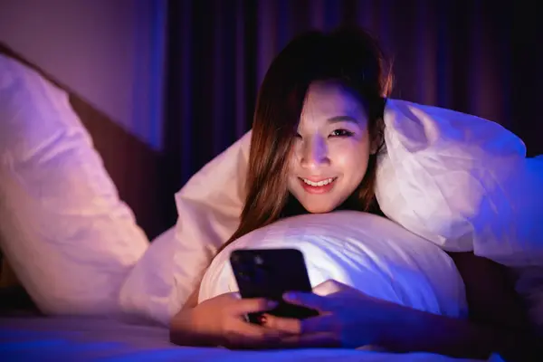Asian woman using mobile phone smartphone laying on the bed in the bedroom. Women scrolling social networks on mobile. Content female lying in bed under blanket and watching movie on smartphone.