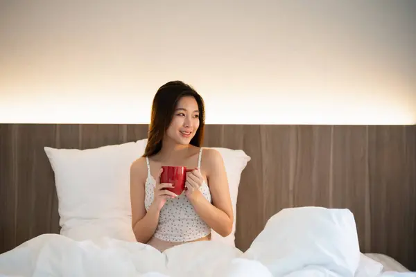 Joyful asian woman drink hot coffee while sitting on the bed in bedroom at home. Asian women relex laying smile after wake up on the bed house. Activity hobby at house concept.
