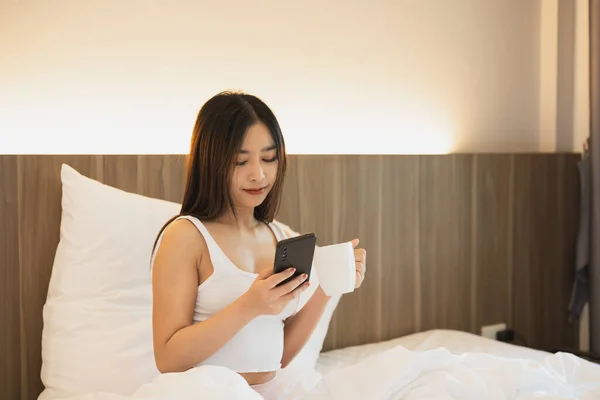 Joyful asian woman drink hot coffee and using mobile phone while sitting on the bed in bedroom at home. Asian women relex laying smile after wake up on the bed house. Activity hobby at house concept.