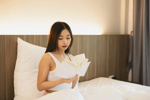 Joyful asian woman drink hot coffee and reading a book while sitting on the bed in bedroom at home. Asian women relax laying smile after wake up on the bed house. Activity hobby at house concept.