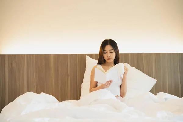 Joyful asian woman drink hot coffee and reading a book while sitting on the bed in bedroom at home. Asian women relax laying smile after wake up on the bed house. Activity hobby at house concept.