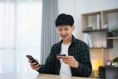 Kredi kartı tutan ve internetten alışveriş yapan bir adam. Evde çalışan Asyalı adam. Çevrimiçi alışveriş, e-ticaret, internet bankacılığı, harcama, ev konseptinden çalışma.
