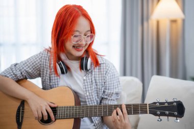 Evdeki oturma odasında kanepede otururken gitar çalan gözlüklü ve kulaklık takan Asyalı bir kadın. Evde gitar çalarken şarkı yazan Asyalı kadınlar. Şarkı müziği konsepti oluştur.