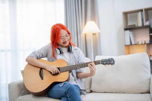 Asian woman wearing glasses and headphone playing guitar while sitting on sofa in the living room at home. Asian women writing song while playing guitar at home. Compose song music concept.