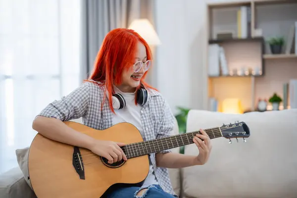 Asian woman wearing glasses and headphone playing guitar while sitting on sofa in the living room at home. Asian women writing song while playing guitar at home. Compose song music concept.