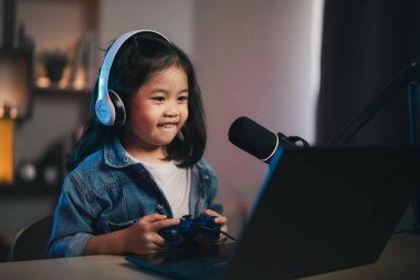 Asyalı etkili genç kız, joystick oynayarak, canlı yayında video konferansını dizüstü bilgisayarla ve kulaklık takarak neşeli bir şekilde gülümsüyor. Çocuklar çevrimiçi oyuncu kadrosu kavramı.
