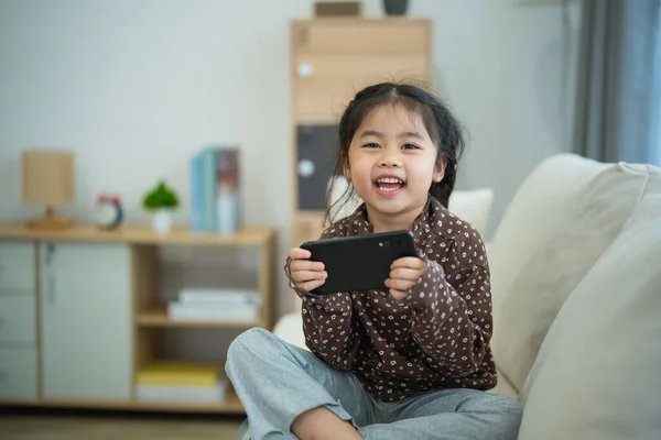 Asian child girl looking using and touch mobile phone screen on couch sofa. Baby smiling funny time to use mobile phone. Too much screen time. Cute girl watching videos while tv, Internet addiction.
