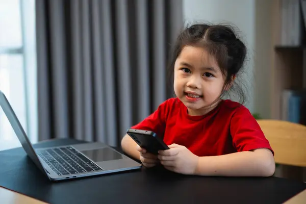 Asian child girl looking using and touch mobile phone screen. Baby smiling funny time to use mobile phone. Too much screen time. Cute girl watching videos while tv in living room, Internet addiction.