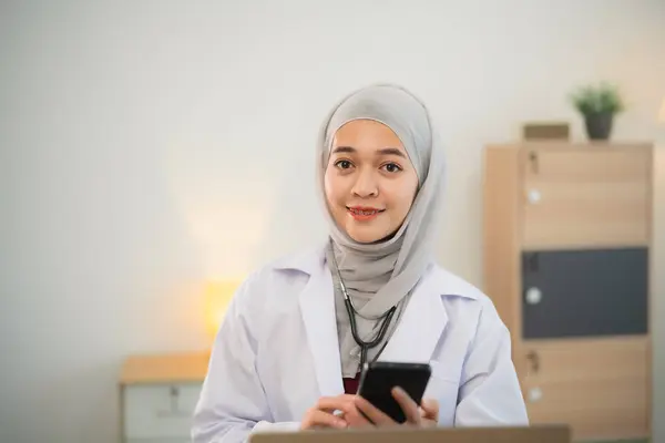 Muslim islam Asian doctor man wearing hijab and stethoscope working with laptop computer and using mobile phone, smartphone. Female doctor work at home office hospital. Health hospital concept.