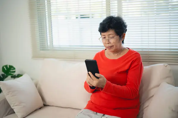 Happy senior old asian woman enjoying using mobile apps texting typing messages sit on sofa, smiling old lady holding smartphone looking smartphone browsing social media or learning technology at home