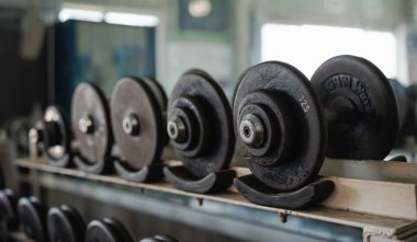 Bir sıra dambıl bir rafa dizilmiştir. Dambıllar siyah ve jimnastik salonunda takılmış gibi görünüyor..