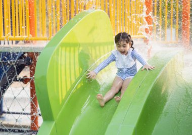 Genç bir kız yeşil bir su kaydırağından kayıyor. Mavi beyaz bir mayo giyiyor ve gülümsüyor.